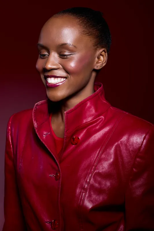Smiling African woman with dark skin wearing a shiny red leather jacket poses against a dark red background. Concept of fashion, beauty, confidence, and elegance in studio lighting.