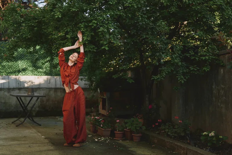 Young woman dancing in a red jumpsuit in a sunlit garden, arms raised in joy, surrounded by lush greenery and a calm outdoor space