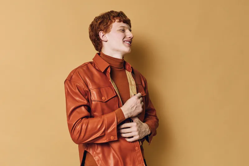 Young man with red hair wearing brown leather jacket and turtleneck sweater standing sideways on beige background with closed eyes and smiling expression.