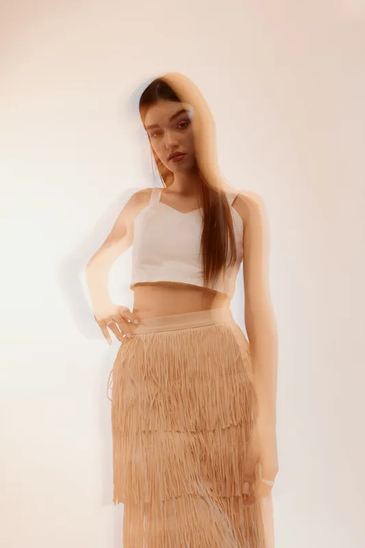 Young woman in a white top and beige fringed skirt poses fashionably with a soft blurred effect. Concept of woman beauty fashion and modern style in studio lighting.