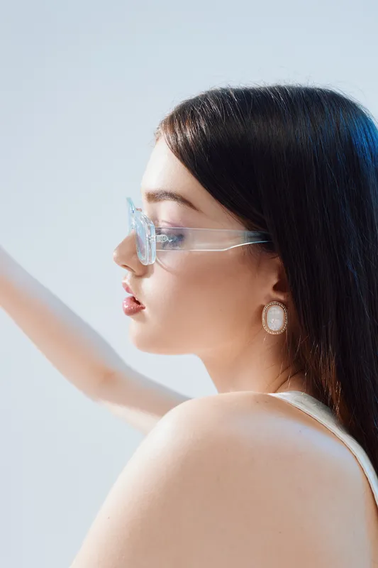 Young woman with long dark hair wearing clear glasses and elegant earrings poses in profile. Soft lighting highlights her smooth skin and beauty. Concept of woman beauty fashion.