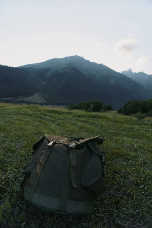 Backpack resting on a grassy meadow with distant mountains and clear sky. Focus on outdoor gear, nature exploration, and rugged solitude in a calm landscape.