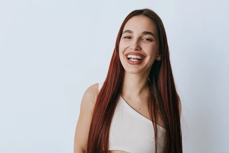 Young woman with long straight hair smiling and looking at camera on white background. Casual clothing and natural makeup with happy expression and relaxed pose in studio.