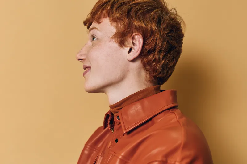Young redhead woman with curly hair wearing brown leather jacket and turtleneck sweater looking sideways against orange background with natural light and profile view.