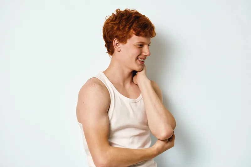 Young man with red hair wearing white tank top smiling and looking sideways. Casual portrait of male model with hand on neck and muscular arms against white background.