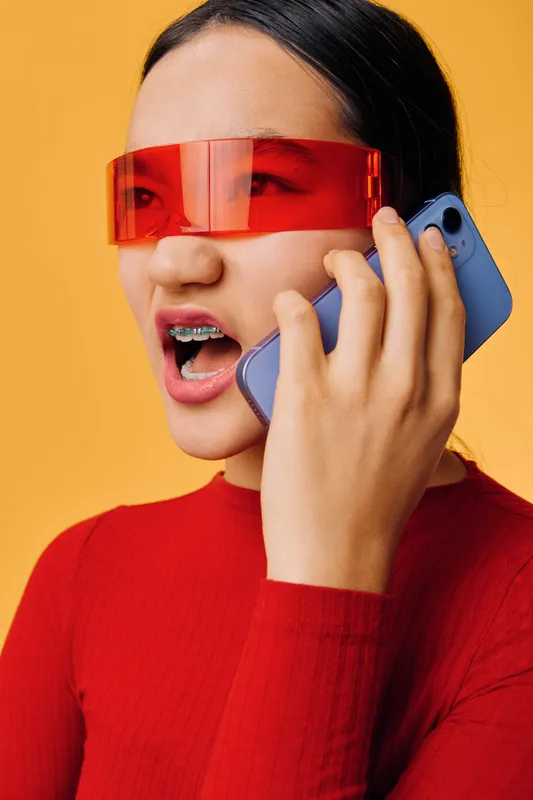 Young woman with braces wearing red sunglasses and red sweater holding blue smartphone talking loudly on orange background for technology communication and style expression.