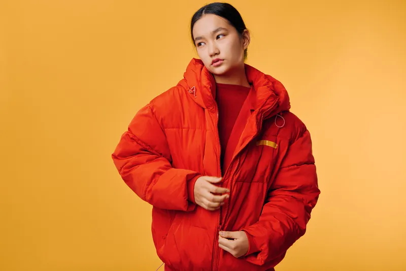 Young woman in bright red puffer jacket against solid yellow background, posing with thoughtful expression and casual style for fashion and winter clothing concept.