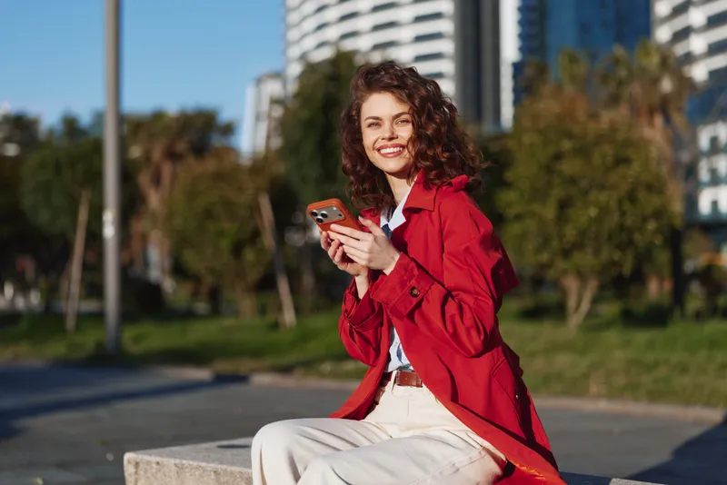 Young woman with curly hair sits on a concrete bench in an urban park, wearing a red jacket and white pants, smiling as she looks at her phone and enjoys a sunny afternoon.