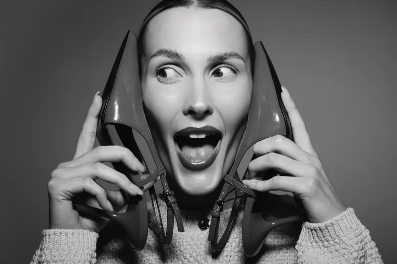 Happy white woman with light skin holds red high heels near face with excited expression on grey background. Concept for fashion, shoes, and joy.