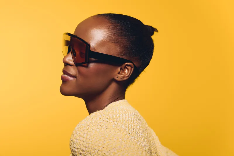 Profile of a confident dark skinned African woman wearing large sunglasses and a yellow knit sweater. Bright yellow background creates a stylish and modern fashion portrait concept.