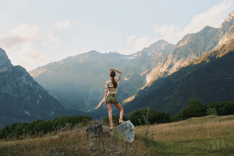 Woman outdoors in casual attire, enjoying a mountain landscape, daylight sun highlights vibrant nature, wide scenic terrain, relaxed lifestyle moment amid rugged slopes