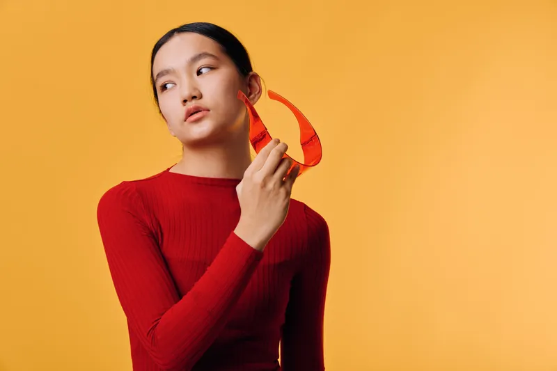Young woman with ponytail and red sweater holding red sunglasses looking to the side on orange background. Studio portrait, fashion, accessory and casual style with confident expression.