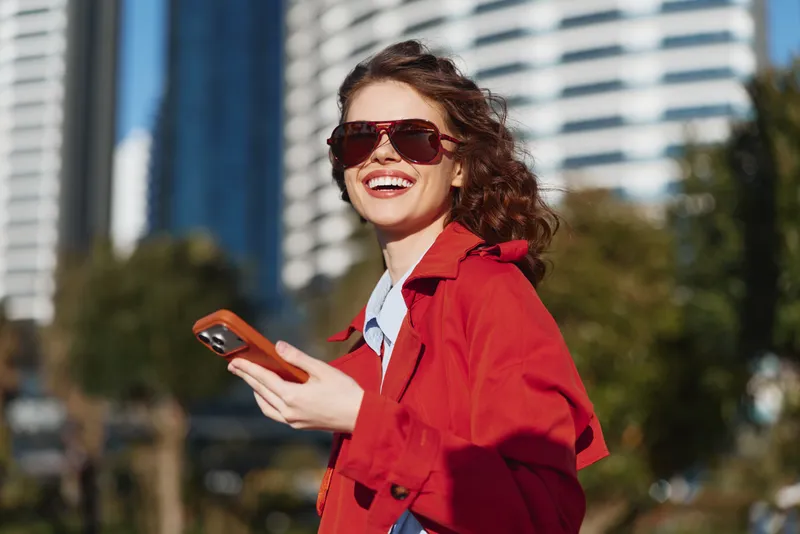 Smiling woman wearing a red coat poses outdoors in an urban park with sunglasses, bright sunlight, and a lively expression, conveying confidence and energy in a modern city setting.