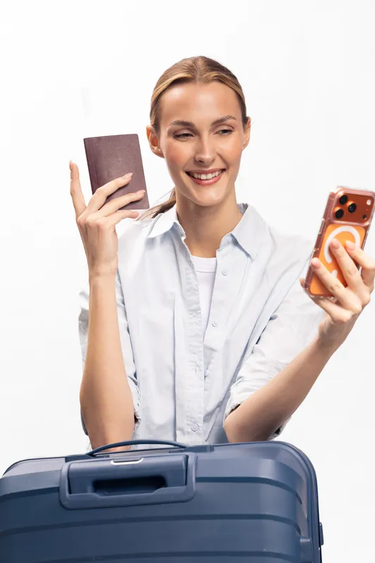 Smiling woman holding passport and smartphone takes selfie with blue suitcase. Travel concept, happy female traveler preparing for trip, isolated on white background in casual shirt and natural light.