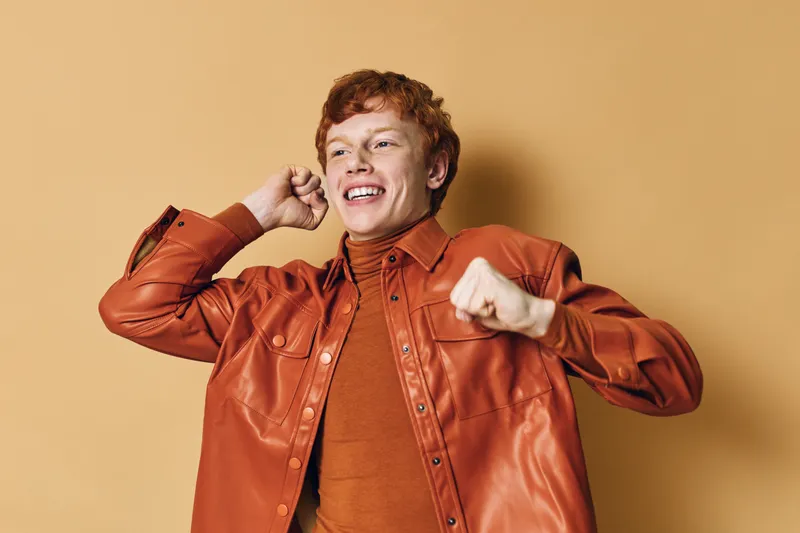 Young man with red hair smiling and dancing wearing an orange leather jacket and turtleneck against beige background. Happy male with joyful expression in casual stylish outfit.