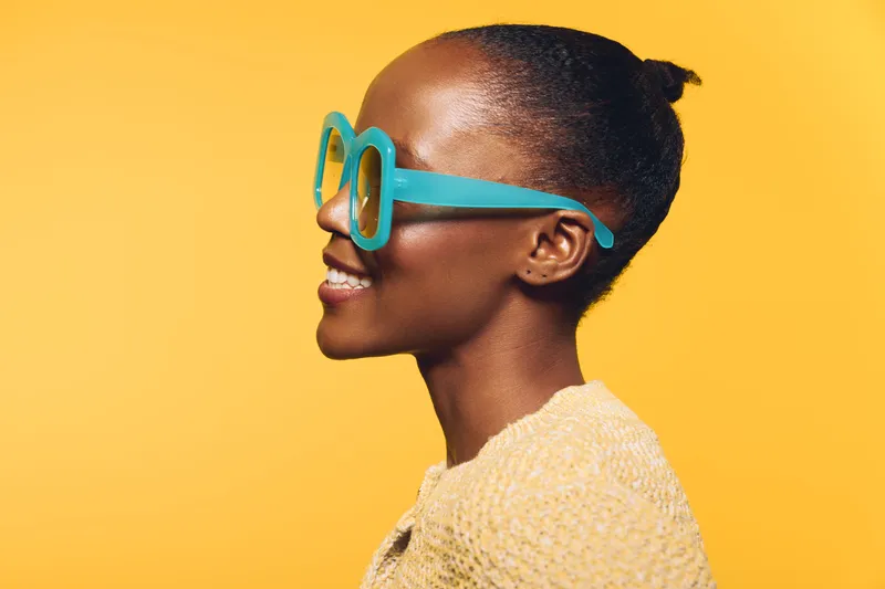 Profile of a smiling African woman with dark skin wearing oversized blue glasses and a textured beige sweater on yellow background. Concept of fashion, style, and confidence.