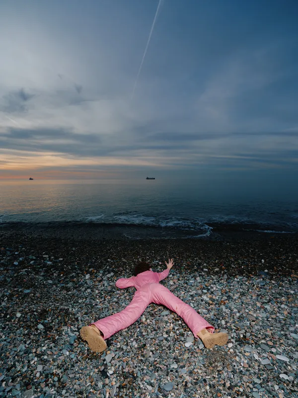 Beach scene with a person in pink on pebbles in a starfish pose, facing the calm sea at sunset, creating a peaceful coastal moment for contemplation.