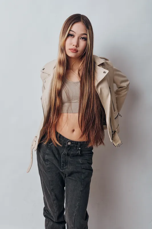 Young woman with long straight hair wearing a beige jacket and crop top posing against light background. Casual style, fashion and confident female with relaxed posture indoors.