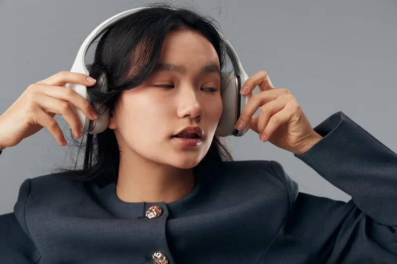 Woman with long black hair and closed eyes wearing headphones and dark jacket listening to music or enjoying sound in studio with plain gray background and relaxed expression.