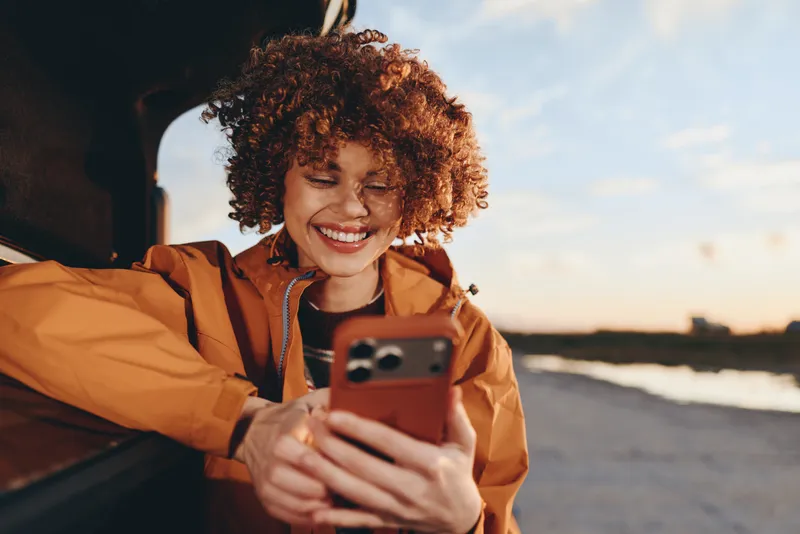 Cheerful woman lifestyle outdoors by coastal shore, curly hair, warm smile, using a smartphone in sunshine, relaxed travel moment near car, golden summer light, soft breeze today