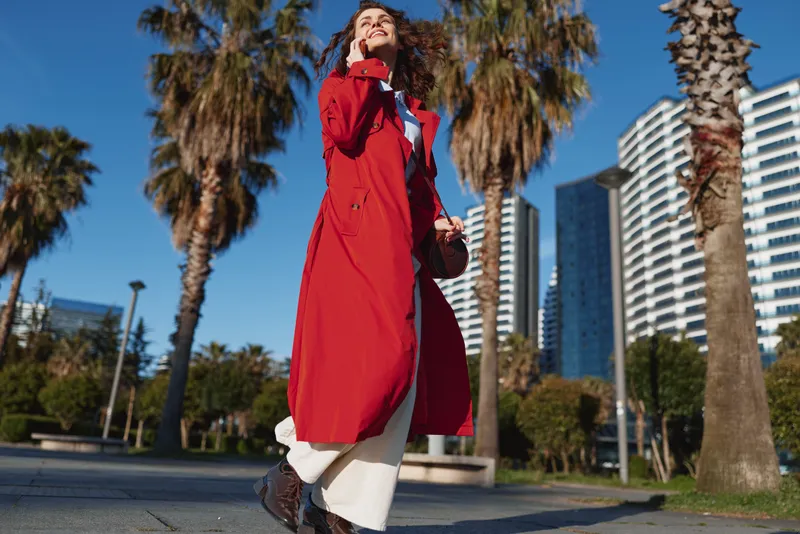 Young woman in a long red coat walks confidently on a sunny urban street lined with palm trees and modern buildings.