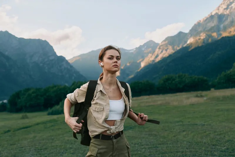 Woman hiking outdoors in a sunlit valley with a backpack, casual jacket and white top, confident pose amid mountains, lush field, and clear blue sky