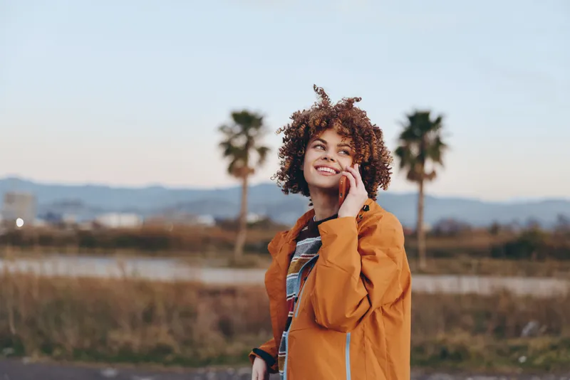 Woman lifestyle outdoors, smiling with curly hair, holding cellphone near ear, orange jacket, standing on rural road, golden autumn light, palm trees blurred, relaxed cheerful mood