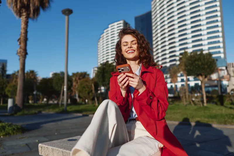 Young woman with curly hair sits on a concrete bench in a city park wearing a red jacket and beige pants while smiling and using her smartphone on a sunny day.