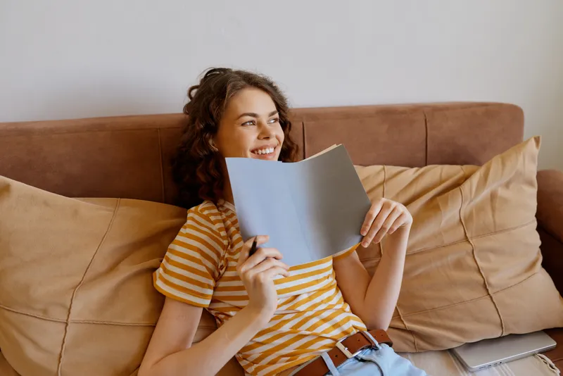 Cheerful woman indoors, lounging on a sofa, reading a notebook, warm sunlight through a window, striped shirt, casual home setting, cozy cushions, relaxed mood, natural colors, everyday moment