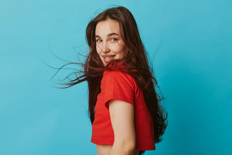 Young woman with long hair wearing red shirt smiling and looking at camera with wind blowing hair on blue background. Portrait of happy female with natural expression and casual style.