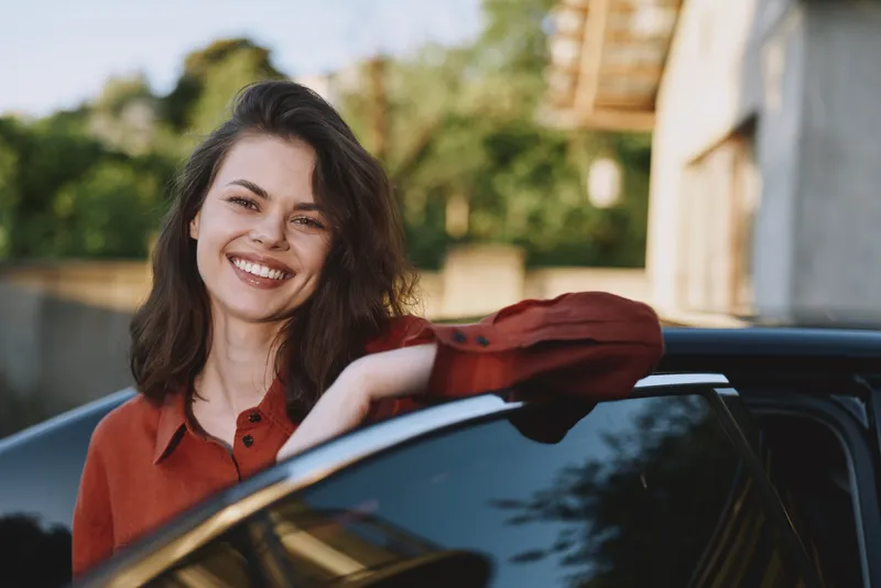 Young woman with shoulder length brunette hair leans on a parked car outdoors, smiling warmly at the camera in a relaxed sunlit moment