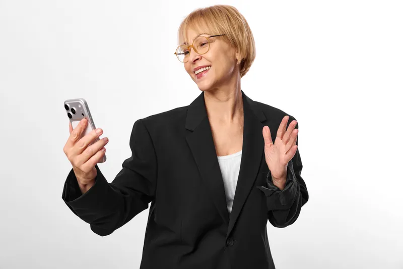 Smiling senior woman with blazer holding a smartphone, making a gesture, studio portrait on white background, communication and technology, cheerful mood