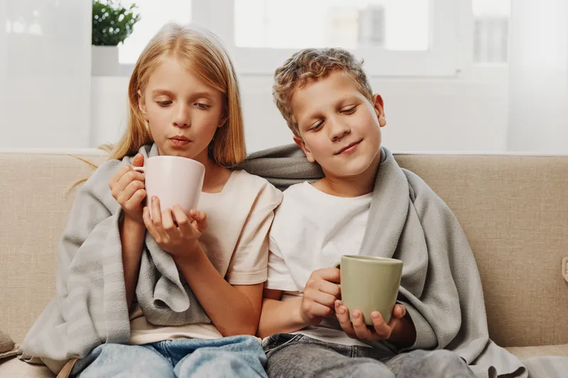 Two children wrapped in a blanket sit on a couch drinking hot beverages, enjoying a cozy moment indoors with soft natural light through the window behind them.