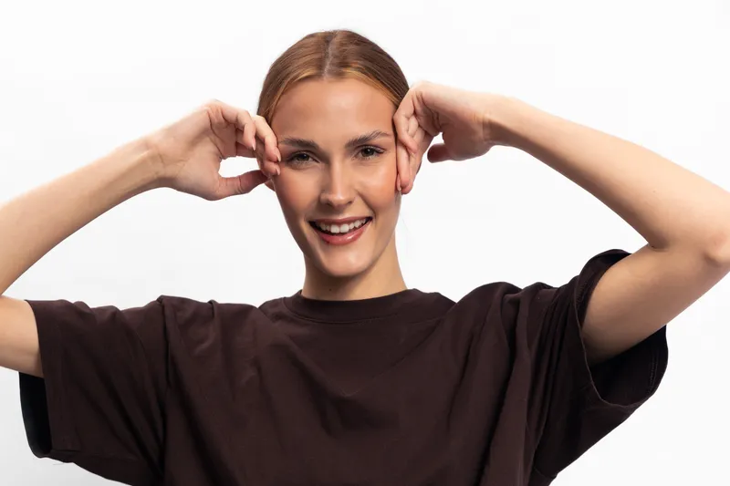 Happy woman with natural skin and pulled back hair smiles gently while posing with hands near face against white background. Concept of happiness, beauty, and natural expression.