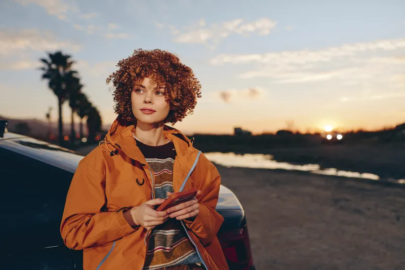 Woman lifestyle portrait outdoors at sunset, curly hair, wearing orange jacket, standing by a car on a seaside road, holding a phone, warm evening light, casual travel mood, soft breeze