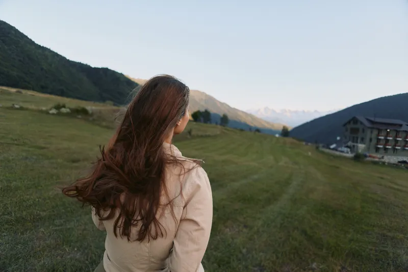 A woman with long brown hair stands outdoors in a green valley surrounded by mountains and buildings, enjoying a peaceful lifestyle moment in nature.