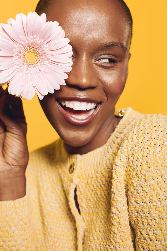 Smiling black woman with dark skin holds pink flower near face, wearing yellow sweater. Bright studio lighting and yellow background create cheerful mood and concept of natural beauty.