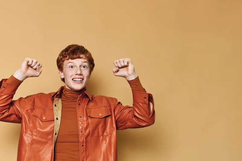 Redhead man with curly hair and leather jacket smiling with both thumbs down gesture on beige background. Young person expressing disapproval, emotion and casual style portrait.