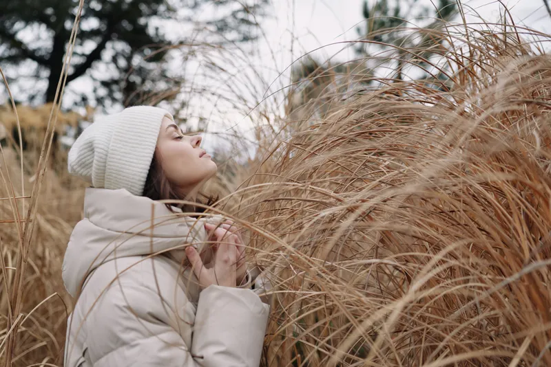 Winter scene featuring a person in a thick coat and knit hat, standing among tall dried grasses, soft light and tranquil mood in a quiet outdoor setting.