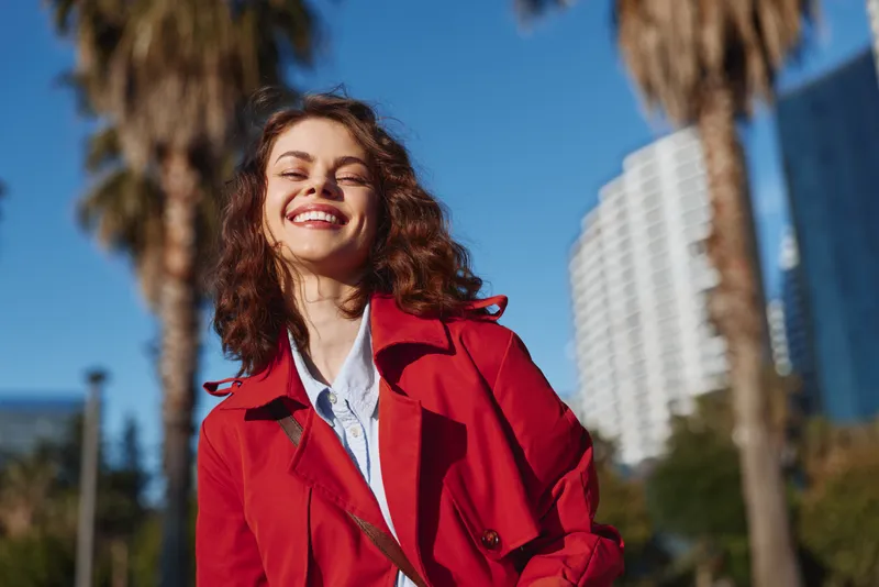 Smiling woman with curly hair wearing red coat outdoors on sunny day, blue sky background, urban environment with palm trees and modern buildings.