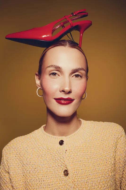 Portrait of a confident woman with red lipstick and hoop earrings balancing a red high heel shoe on her head against a warm brown background. Concept of fashion and creativity.
