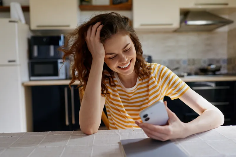 Young woman with wavy hair sits at a bright kitchen table, smiling as she looks at her smartphone, enjoying a casual moment of connection and everyday life in a cozy home