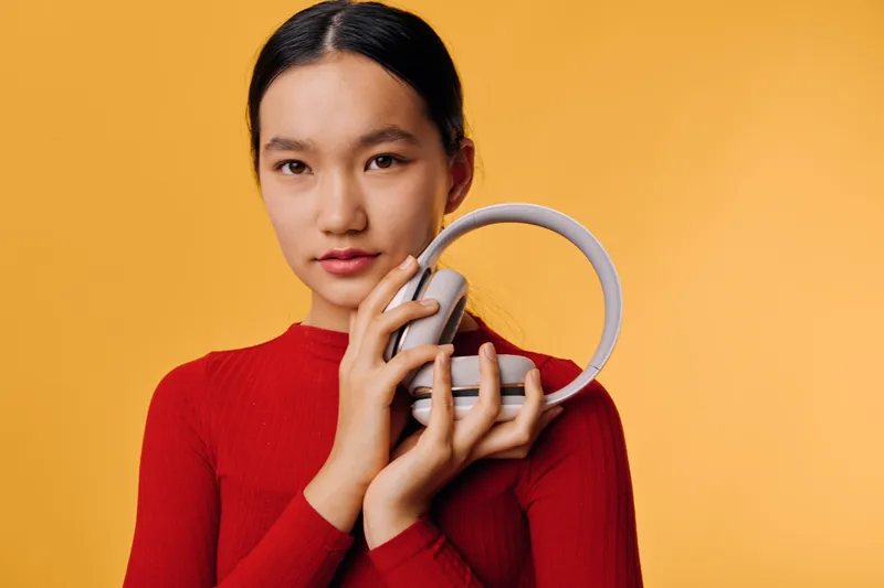 Young woman holding headphones close to face on yellow background for music listening, technology, lifestyle, and relaxation with casual red outfit and natural expression.