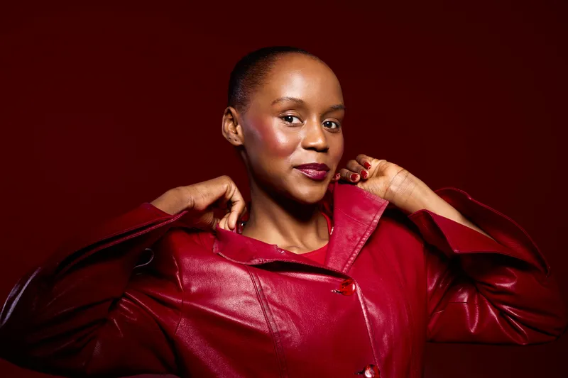 Confident African American woman with dark skin wearing a red leather jacket poses against a red background. Studio portrait highlights fashion and elegance concept.
