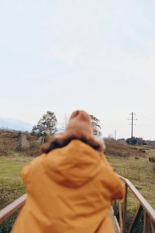 Woman lifestyle hiking in countryside outdoors, back view of person, wearing yellow hood jacket, walking on wooden bridge rail, facing road ahead, calm travel moment, natural daylight, soft blur