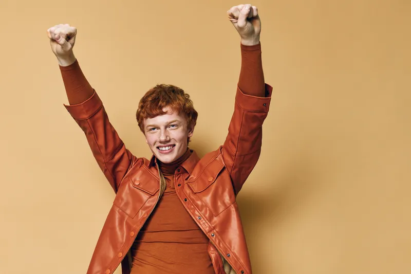 Young man with red hair celebrating success with raised fists and smile wearing orange leather jacket and sweater on beige background with joyful expression.