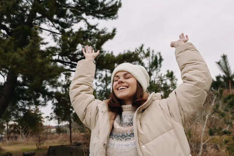 Woman in a puffy winter coat and beanie raises arms in joy outdoors, smiling under trees on a chilly windy day.