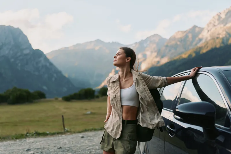 Young woman lifestyle traveler leans on car outdoors in mountains enjoys fresh air and freedom casual outfit summer day scenic nature background peaceful moment