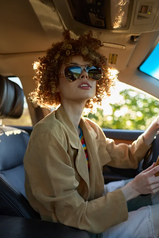 Cheerful woman driving, wearing sunglasses, curly hair, sunlit interior, retro film color vibe, casual jacket, relaxed posture, warm tones, natural light, vibrant scene in a car setting