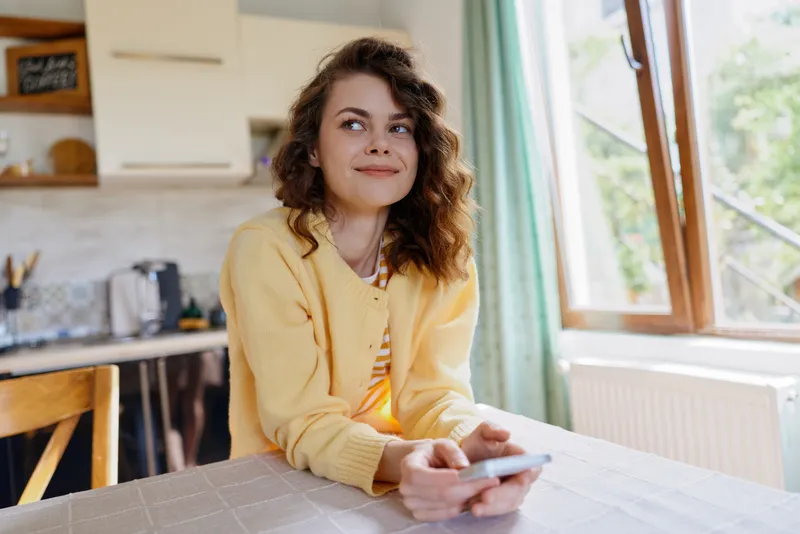 Cheerful woman indoors, seated at a table, wearing a yellow cardigan, holding a phone, sunlight through a window, bright cozy home lifestyle moment, natural colors